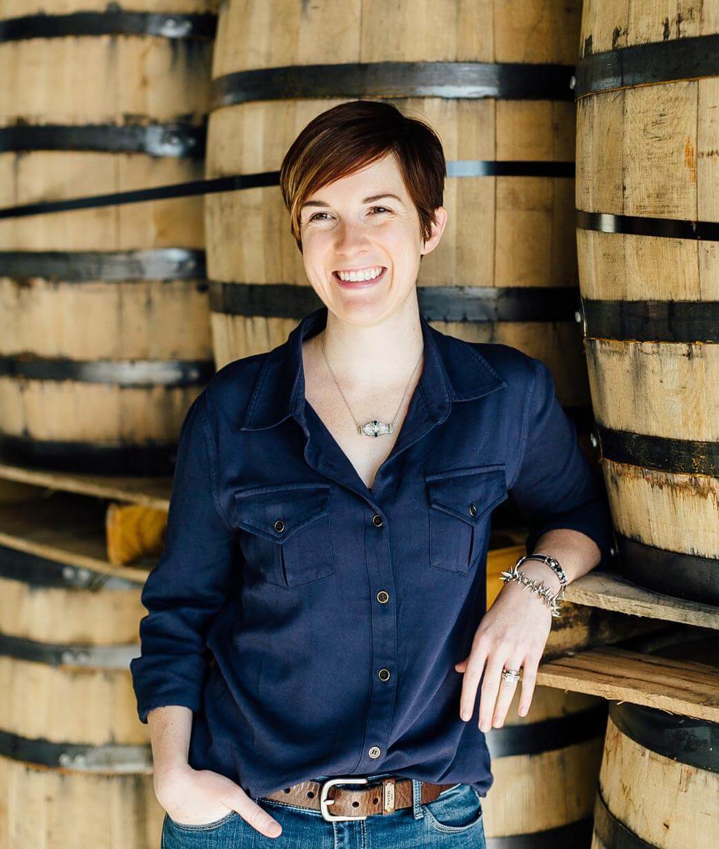 Master Distiller Alex Castle leaning on whiskey barrels and smiling