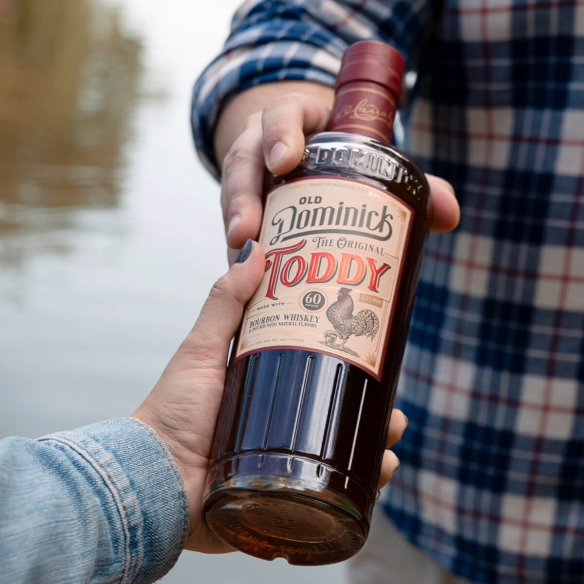 Person in plaid shirt handing bottle of Old Dominick Original Toddy to person wearing a jean jacket, in front of a lake